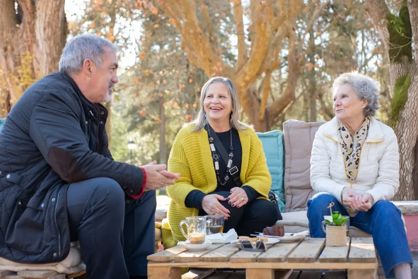 Three older friends laughing together at a garden table.