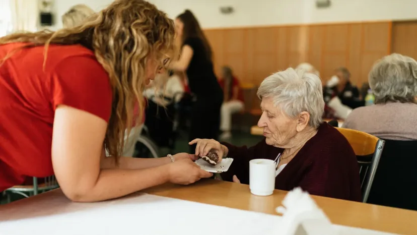 A carer leaning in to chat with an older woman at a dining table.